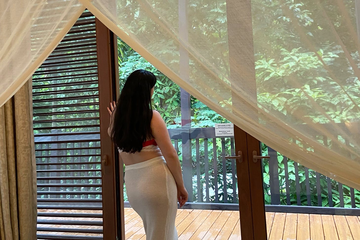 Woman in a hotel staring out the window to a garden.